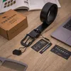 A black leather belt is laid out on a wooden surface next to its packaging, tags, and a laptop. Produktbild-Vorschau
