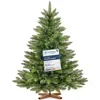 A green artificial Christmas tree with a wooden stand and a tag attached to one of its branches. Produktbild-Vorschau