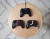 Three black Nintendo Switch controllers are arranged on a round wooden table, with a white and gray patterned rug visible in the background. Anteprima dell’immagine prodotto