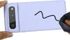 A hand holds a light purple phone case, showing the camera area and a black squiggle drawn on the back. Another hand is wiping the case with a dark cloth. Produktbild-Vorschau