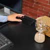 A person's hand is interacting with a cable management accessory on a black desk, with a computer monitor in the background and a brick wall behind it. An hourglass is also visible on the desk. Náhled obrázku produktu
