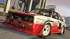 A rally car with racing decals is shown driving on a city street. Produktbild-Vorschau