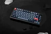 A keyboard is placed on a dark, wet surface, surrounded by black sand and rocks. Produktbild-Vorschau