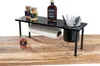 A black metal rack on a wooden surface, holding spices, barbecue sauce, paper towels, and cutlery. The background is white. Náhled obrázku produktu