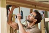 A man is using a Bosch cordless drill to attach a wooden beam to a larger wooden structure. He is wearing safety glasses and appears to be in a workshop setting. Náhled obrázku produktu