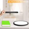 A hand holding a thin black object, with a green book and a round white object on a table below. The background is blurred. Voir les images du produit