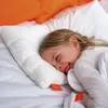 A young girl is happily lying on a white pillow on a bed, with orange pillows in the background. Produktbild-Vorschau