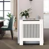 A white radiator cover with vertical slats is placed against a light pink wall. A bouquet of flowers in a white vase sits on top of the cover. To the left, a dining table and chairs are visible. Náhled obrázku produktu
