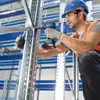 A worker in a warehouse is using a Bosch cordless drill to assemble metal shelving. Produktbild-Vorschau