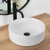 A white ceramic basin with a ribbed exterior sits on a wooden countertop, with a black faucet and a round mirror in the background. Podgląd zdjęcia produktu
