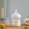 A baby bottle filled with milk sits on a wooden surface, with baby toys and a folded towel in the background. Náhľad obrázku produktu