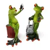 Two decorative frog figurines, one sitting on a red suitcase and taking a selfie with a phone, the other standing with a silver suitcase. Produktbild-Vorschau