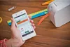 A hand holding a smartphone and a label printer on a wooden desk with office supplies. Produktbild-Vorschau