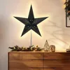 A black star-shaped lamp with small gold star cutouts is displayed on a wooden dresser, illuminated by a warm light. Decorative greenery with fairy lights is placed in front of the lamp, and two geometric lanterns are to the right. Produktbild-Vorschau