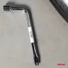 A silver L-shaped lug wrench with a white label is placed on a light gray concrete surface. A portion of a tire is visible in the upper right corner. Náhľad obrázku produktu