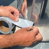 A close-up of hands applying grey tape to a silver cylindrical object. Produktbild-Vorschau