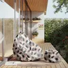 A beanbag chair and ottoman with a feather pattern are placed on a wooden deck next to a glass-enclosed room and a lush green hedge. Náhled obrázku produktu