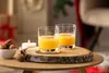 Two glasses of orange juice are placed on a wooden slice on a table, with walnuts and a candle in the background. Produktbild-Vorschau