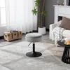 A gray upholstered stool with a black base sits on a patterned rug in a living room setting. A wooden chest and a plant are visible in the background. Produktbild-Vorschau