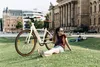 A woman sits on a grassy lawn next to a white e-bike, with a large building in the background. Produktbild-Vorschau