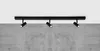 A black track light fixture with three adjustable spotlights mounted on a ceiling above a concrete wall. Náhľad obrázku produktu