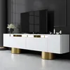 A modern white TV stand with gold accents and cylindrical gold legs, featuring a large black television on top. Náhľad obrázku produktu