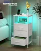 A white nightstand with drawers and an open shelf, featuring a phone charging, a radio, and a vase with flowers. The nightstand has integrated LED lighting. Produktbild-Vorschau