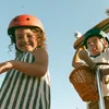 Two children wearing helmets, one with a skateboard, the other with a scooter. Produktbild-Vorschau