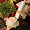 A hand holding a portable cake printer over a cup of coffee with Christmas-themed latte art, surrounded by Christmas gifts and decorations. Podgląd zdjęcia produktu