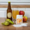A still life arrangement of cider-making ingredients, including a bottle of cider, a glass of cider, apples, pears, and a packet of yeast on a wooden surface. Produktbild-Vorschau