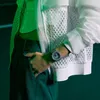 A person's arm wearing a watch is shown in a close-up shot, with a green and white outfit and a dark green background. Produktbild-Vorschau