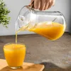 A hand is pouring orange juice from a glass pitcher into a glass on a wooden board. Podgląd zdjęcia produktu