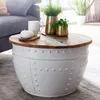 A round coffee table with a wooden top and a white riveted base, placed in a living room setting with a sofa and plants. Produktbild-Vorschau