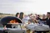 People toasting drinks around a table with a pizza oven in the foreground. Produktbild-Vorschau