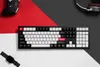 A black mechanical keyboard is displayed on a surface with red and gray sections, alongside a black game controller and brown headphones. Produktbild-Vorschau