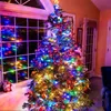A decorated Christmas tree with colorful lights in a room, next to a window with lights reflecting. Náhled obrázku produktu