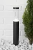 A modern black and clear bollard light is placed on white stones in front of a white brick wall and a green bush. Produktbild-Vorschau