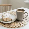 A close-up of a ceramic mug filled with coffee and a cinnamon roll on a plate, both placed on a woven placemat on a white table. Produktbild-Vorschau