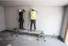 Two construction workers are installing drywall on a wall while standing on a portable platform. Produktbild-Vorschau