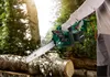 A person is using an electric chainsaw to cut a log in a forest setting. Produktbild-Vorschau