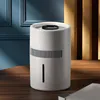 A white humidifier/air purifier sits on a wooden table in front of a blue wall and a textured yellow partition. Náhled obrázku produktu