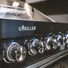 Close-up of the control panel of a black gas grill, showing several chrome knobs and the brand logo. Náhled obrázku produktu