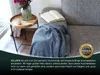 A blue blanket is draped over a gray sofa next to a small wooden side table with a vase of flowers and a book. Produktbild-Vorschau
