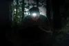 A man wearing a headlamp is crouching in a dark forest, looking at his phone. Podgląd zdjęcia produktu