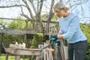 A woman is using a cordless chainsaw in a garden setting, with a treehouse in the background. Produktbild-Vorschau