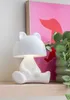 A white bear-shaped table lamp is placed on a shelf next to a potted plant and a framed pink artwork. Náhled obrázku produktu