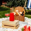 A picnic scene with a wooden crate, a carafe of red juice, a glass, a picnic basket with flowers and a bottle, a book, and watermelon slices on a blanket. Produktbild-Vorschau