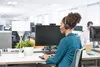 A woman wearing a headset works at a computer in an office setting. Anteprima dell’immagine prodotto