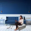A woman is sitting on a folded mattress in a living room, using a laptop. Voir les images du produit