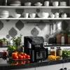 A black air fryer sits on a kitchen counter with fresh vegetables and spices. Produktbild-Vorschau